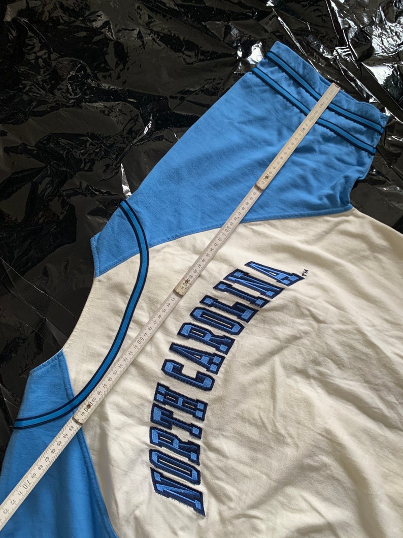 May include: A white and light blue baseball jersey with the text "NORTH CAROLINA" embroidered in blue on the front. A measuring tape is visible in the image.