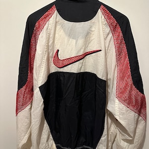 Könnte beinhalten: Eine schwarze, weiße und rote Windbreaker-Jacke mit einem großen Nike Swoosh-Logo auf der Rückseite. Die Jacke hat Mesh-Einsätze an den Seiten und einen Reißverschluss.