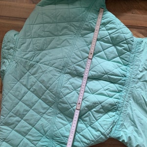 May include: A light blue quilted jacket with a diamond pattern. The jacket is laid flat and a measuring tape is placed on top of it.