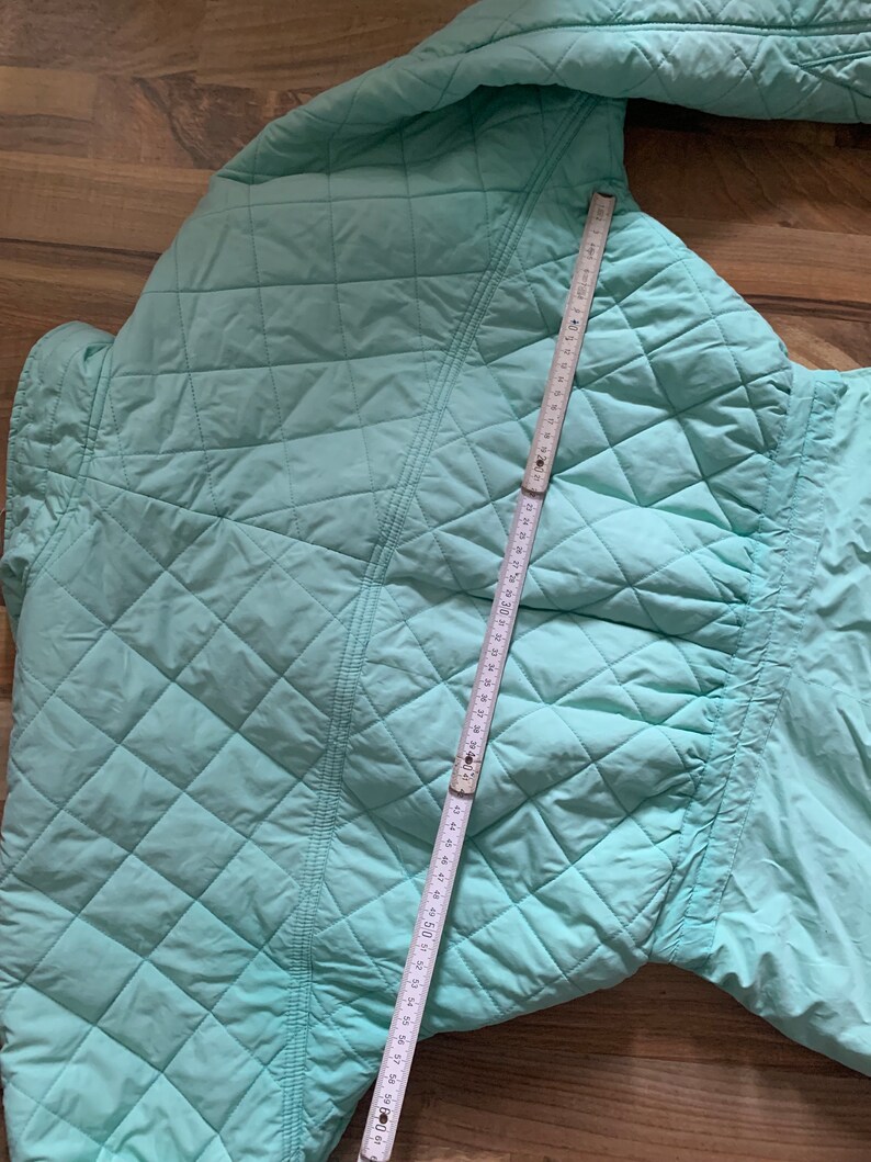 May include: A light blue quilted jacket with a diamond pattern. The jacket is laid flat and a measuring tape is placed on top of it.