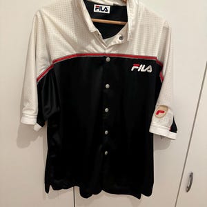 Peut inclure: Chemise boutonn&eacute;e avec une partie sup&eacute;rieure en maille blanche et une partie inf&eacute;rieure noire. La chemise a une bande rouge sur la poitrine et le logo Fila est visible. Les manches sont aux trois quarts.
