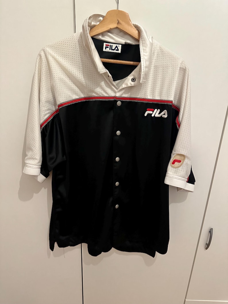 Peut inclure: Chemise boutonn&eacute;e avec une partie sup&eacute;rieure en maille blanche et une partie inf&eacute;rieure noire. La chemise a une bande rouge sur la poitrine et le logo Fila est visible. Les manches sont aux trois quarts.