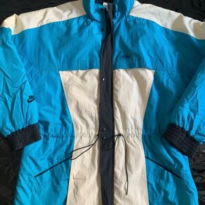 May include: A vintage Nike ski jacket in blue, white, and black with a drawstring waist and a zipper closure. The jacket has a Nike logo on the left sleeve and a Nike text logo on the right chest.