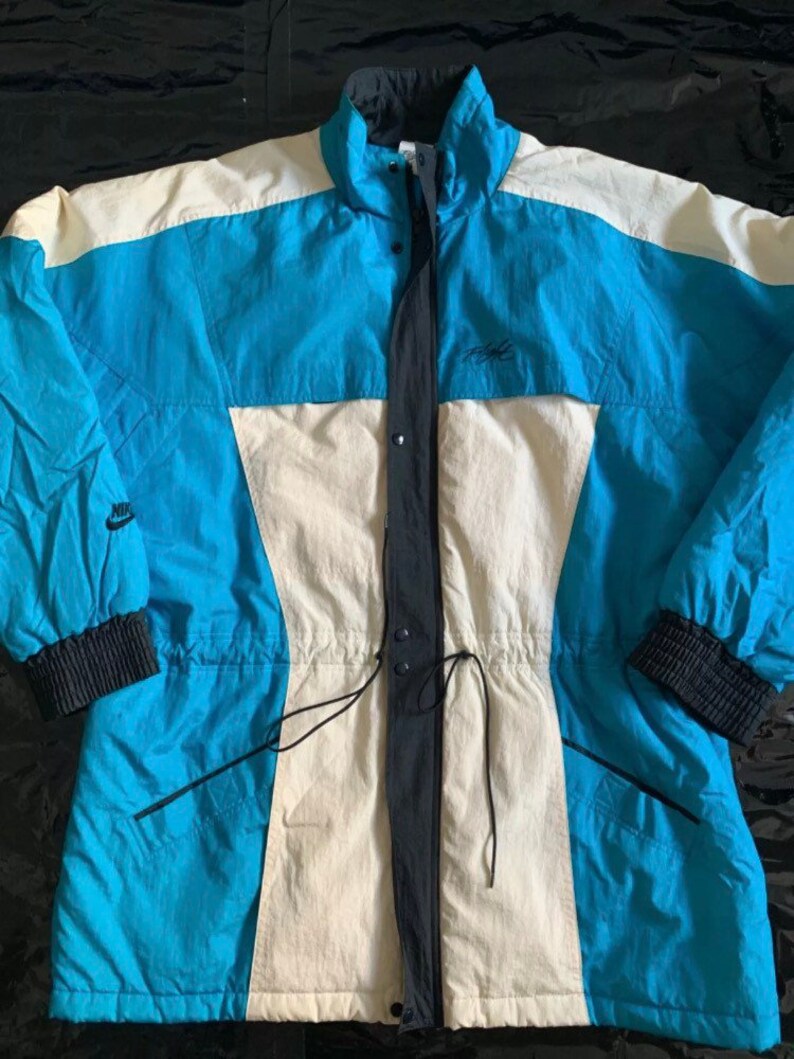 May include: A vintage Nike ski jacket in blue, white, and black with a drawstring waist and a zipper closure. The jacket has a Nike logo on the left sleeve and a Nike text logo on the right chest.