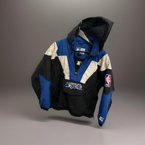 Op de afbeelding: Een zwarte, blauwe en witte Starter anorakjas met capuchon. De jas heeft het Orlando Magic logo en een NBA-patch. De jas heeft een halve ritssluiting en een Starter logo op de mouw.