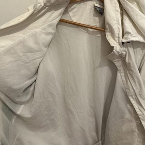 Puede incluir: Chaqueta con capucha de color claro con forro de malla, colgada de una percha de madera. La chaqueta est&aacute; abierta, revelando el forro interior y el cord&oacute;n en la parte inferior. La chaqueta parece estar hecha de un material ligero.