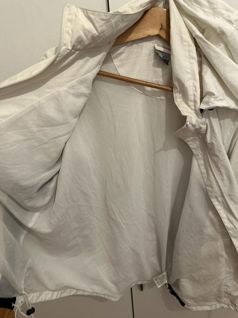 Puede incluir: Chaqueta con capucha de color claro con forro de malla, colgada de una percha de madera. La chaqueta est&aacute; abierta, revelando el forro interior y el cord&oacute;n en la parte inferior. La chaqueta parece estar hecha de un material ligero.