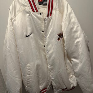 Op de afbeelding: Witte satijnen baseballjas met rood-witte streepjes aan de zoom. De jas heeft een drukknoopsluiting en een Nike-logo op de linkerborst. De achterkant van de jas heeft een ster met de tekst "76ers" erop. De jas is maat 3XL.