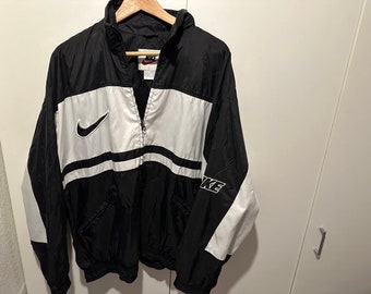 Könnte beinhalten: Schwarze und weiße Windbreaker-Jacke mit einem weißen Nike Swoosh-Logo auf der linken Brust und dem Wort "NIKE" auf dem rechten Ärmel.