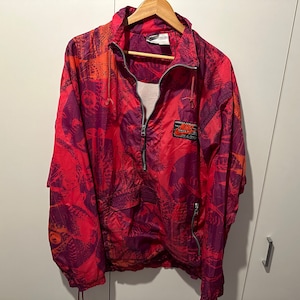 Puede incluir: Una chaqueta cortavientos roja y morada con un logotipo de Nike en el pecho izquierdo. La chaqueta tiene una cremallera y una capucha con cordón.