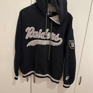 Könnte beinhalten: Schwarzer Raiders-Hoodie mit dem Teamnamen in weißer, stilisierter Schrift auf der Brust. Der Hoodie hat eine Kapuze mit Kordelzug, einen Teamlogo-Aufnäher auf dem Ärmel und einen weiß-grauen Streifen.