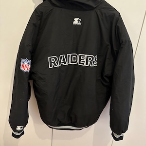 Könnte beinhalten: Schwarze Raiders-Jacke von Starter mit Kapuze. Das Wort "RAIDERS" ist weiß auf dem Rücken aufgedruckt. Ein NFL-Patch befindet sich auf dem linken Ärmel. Die Jacke hat einen weiß-schwarzen Streifen. Das Starter-Logo ist sichtbar.