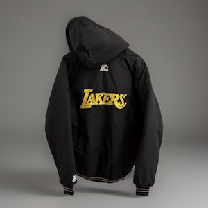 Puede incluir: Chaqueta negra con capucha con la palabra "Lakers" en letras doradas en la espalda. La chaqueta tiene un logotipo de Starter en la capucha y puños con rayas rojas, blancas y moradas.
