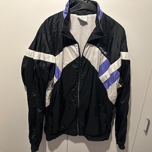 Könnte beinhalten: Schwarze Windbreaker-Jacke mit weißen und lila Streifen. Die Jacke hat einen Reißverschluss und einen Stehkragen.