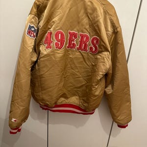 Könnte beinhalten: Eine goldene Satin-Bomberjacke mit dem roten, gestickten Schriftzug 49ERS auf der Rückseite. Die Jacke hat einen rot-weiß gestreiften Kragen, Bündchen und Saum sowie einen NFL-Logo-Aufnäher am rechten Ärmel und ein Starter-Logo am linken Ärmel.