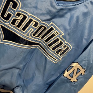 K&ouml;nnte beinhalten: Eine hellblaue Jacke mit einem wei&szlig;en und marineblauen "Carolina"-Logo auf der Vorderseite und einem wei&szlig;en und marineblauen "N.C." -Logo auf dem &Auml;rmel.