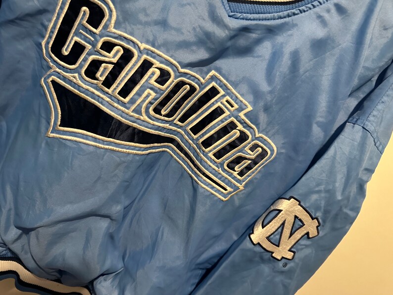K&ouml;nnte beinhalten: Eine hellblaue Jacke mit einem wei&szlig;en und marineblauen "Carolina"-Logo auf der Vorderseite und einem wei&szlig;en und marineblauen "N.C." -Logo auf dem &Auml;rmel.