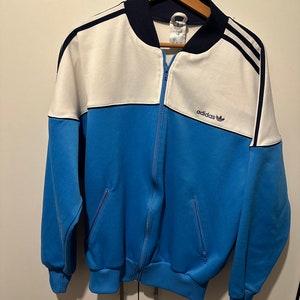 Op de afbeelding: Een vintage Adidas trainingsjack in blauw, wit en donkerblauw met een ritssluiting en het Adidas logo op de voorkant.