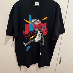 May include: Black Nike t-shirt with a basketball player dunking. The design features the words "JAM" and "FREQUENCY" in red and blue, with lightning bolt accents. The player wears a white jersey with the number 23.