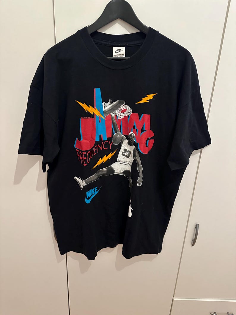 May include: Black Nike t-shirt with a basketball player dunking. The design features the words "JAM" and "FREQUENCY" in red and blue, with lightning bolt accents. The player wears a white jersey with the number 23.