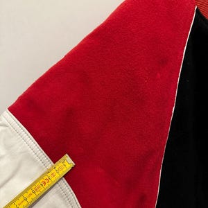 May include: Close-up of a varsity jacket sleeve. The sleeve features a red felt panel, a white leather panel, and a black panel. The red panel is bordered by white piping. A yellow ruler is placed across the white leather panel.