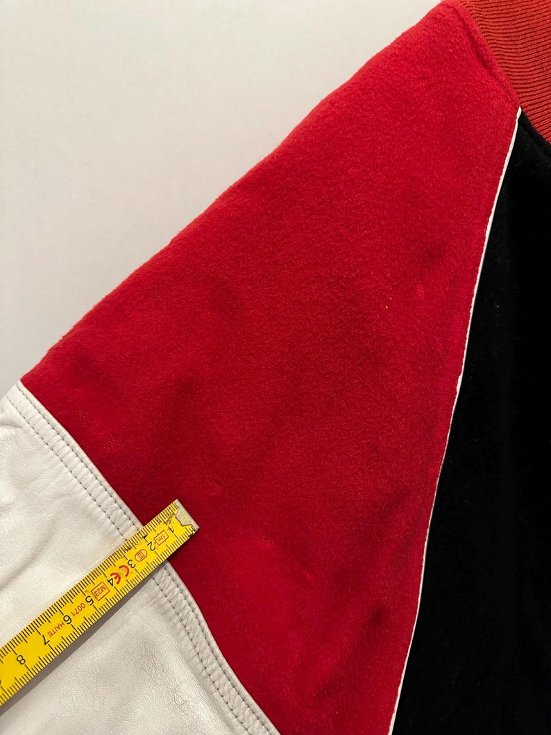 May include: Close-up of a varsity jacket sleeve. The sleeve features a red felt panel, a white leather panel, and a black panel. The red panel is bordered by white piping. A yellow ruler is placed across the white leather panel.