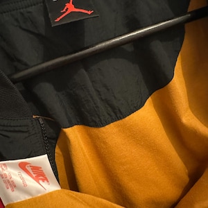 K&ouml;nnte beinhalten: Schwarze und goldene Nike Jacke mit rotem Jumpman-Logo. Die Jacke hat einen schwarzen oberen Teil und einen goldenen unteren Teil. Das Nike-Logo ist auf einem Etikett sichtbar. Gr&ouml;&szlig;e XL.