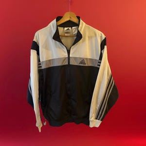 May include: A black and white Adidas track jacket with a zipper closure and three white stripes on each sleeve. The jacket has a silver reflective stripe across the chest and the Adidas logo is on the left chest.