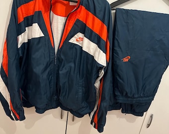 May include: A navy blue Nike tracksuit, with a jacket featuring orange and white stripes and matching trousers. The jacket has a full zip and the Nike logo is visible on both items. The tracksuit is hanging on a black hanger.