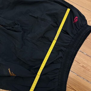 K&ouml;nnte beinhalten: Schwarze Sporthose mit goldenem Logo und rotem Nike-Logo. Die Hose hat einen elastischen Bund und wird mit einem gelben Lineal gemessen. Die Hose ist aus leichtem Material.