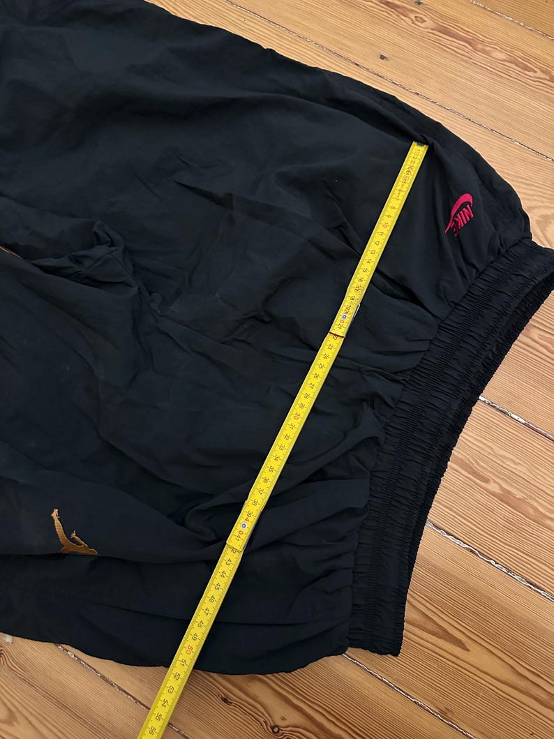 K&ouml;nnte beinhalten: Schwarze Sporthose mit goldenem Logo und rotem Nike-Logo. Die Hose hat einen elastischen Bund und wird mit einem gelben Lineal gemessen. Die Hose ist aus leichtem Material.