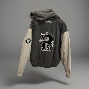 Könnte beinhalten: Grau-beige Raiders-Jacke mit Kapuze. Auf der Rückseite befindet sich ein großes "P"-Design mit dem Teamlogo und dem Wort "Raiders". Der linke Ärmel hat einen Raiders-Logo-Aufnäher. Die Jacke ist von der Marke Starter.