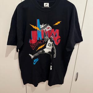 May include: Black Nike t-shirt with a basketball player graphic dunking. The design features the words "JAMM" and "FREQUENCY" in red and blue, with lightning bolt accents. The Nike logo is visible on the shirt.