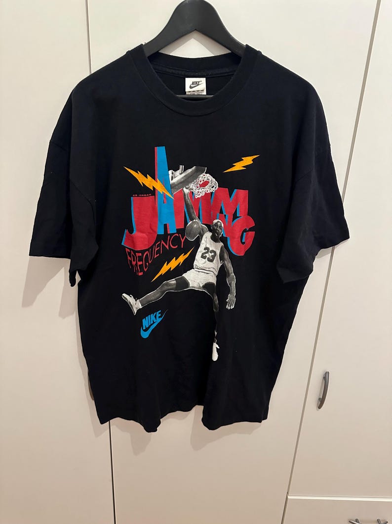 May include: Black Nike t-shirt with a basketball player graphic dunking. The design features the words "JAMM" and "FREQUENCY" in red and blue, with lightning bolt accents. The Nike logo is visible on the shirt.