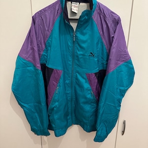 Könnte beinhalten: Eine Vintage Puma Trainingsjacke in Teal, Lila und Schwarz. Die Jacke hat einen durchgehenden Reißverschluss, einen Stehkragen und zwei Reißverschlusstaschen. Das Puma-Logo ist auf der Brust sichtbar. Die Jacke hängt an einem schwarzen Kleiderbügel.