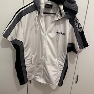 Op de afbeelding: Witte hoodie met korte mouwen en ritssluiting met zwarte en grijze accenten. Voorzien van het Adidas-logo op de mouw en borst. De hoodie heeft een trekkoord en twee voorzakken. De capuchon is grijs en zwart.