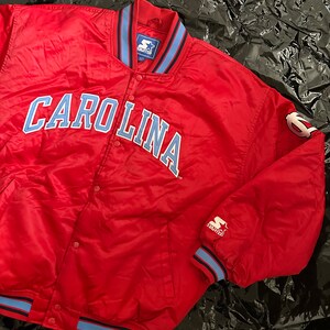 May include: Red satin bomber jacket with blue and black trim. The jacket features the word "CAROLINA" in blue on the front and a Starter logo on the sleeve.
