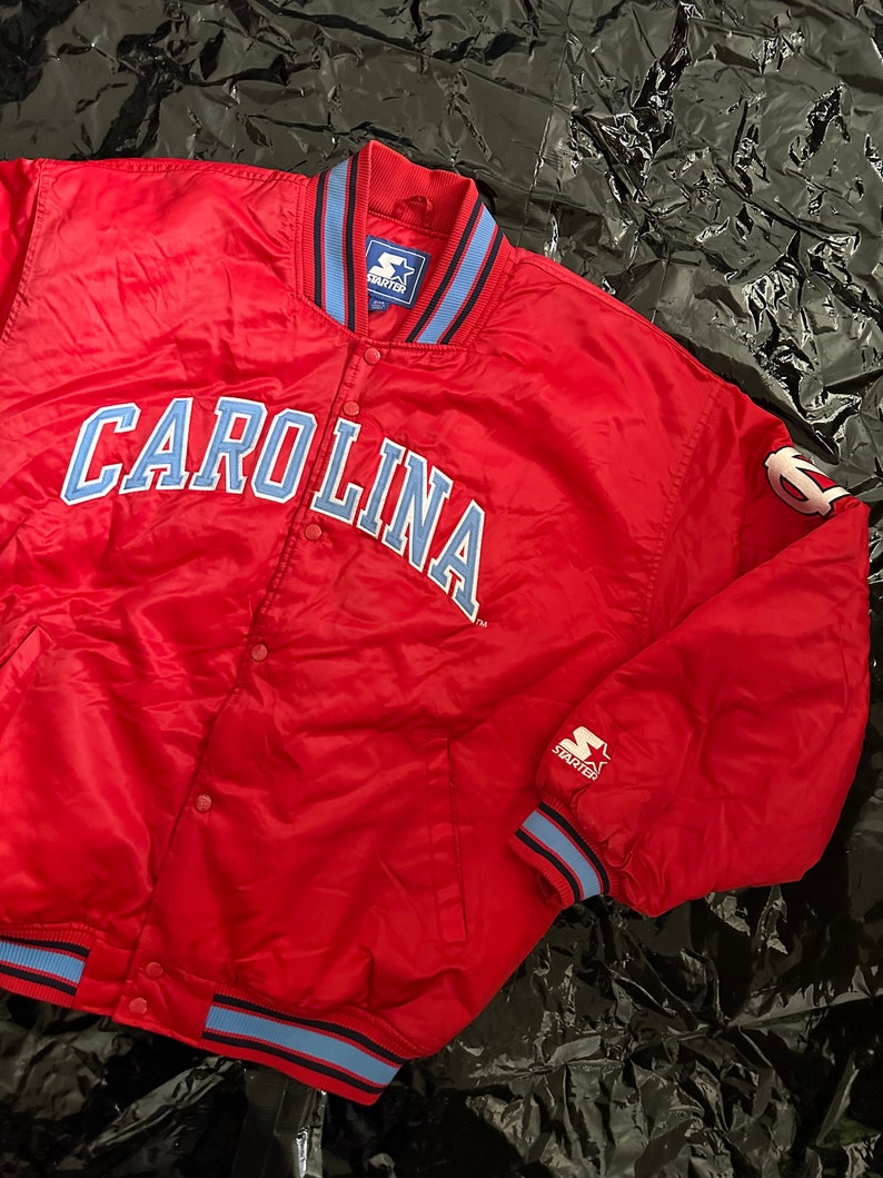 May include: Red satin bomber jacket with blue and black trim. The jacket features the word "CAROLINA" in blue on the front and a Starter logo on the sleeve.