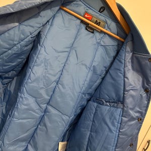May include: Light blue varsity jacket with a quilted interior. The jacket features a ribbed collar and cuffs with white stripes. The Nike logo is visible on the label. The jacket is hanging on a wooden hanger.