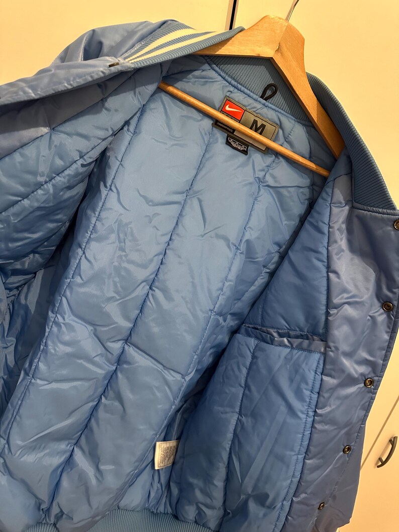 May include: Light blue varsity jacket with a quilted interior. The jacket features a ribbed collar and cuffs with white stripes. The Nike logo is visible on the label. The jacket is hanging on a wooden hanger.
