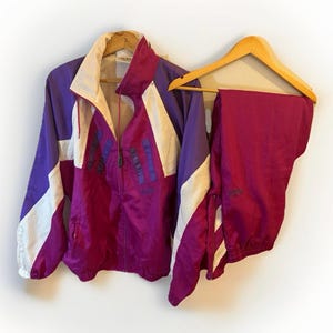May include: A vintage purple, white, and magenta tracksuit. The jacket has a zip-up front, a high collar, and white accents. The matching pants are a solid magenta color. Both pieces are hanging on wooden hangers.