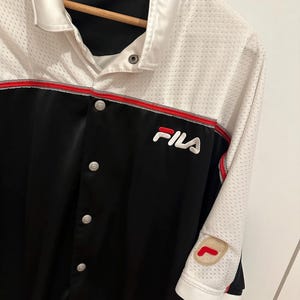 Peut inclure: Chemise boutonn&eacute;e Fila noire et blanche avec garniture rouge. La chemise pr&eacute;sente le logo Fila sur la poitrine et un &eacute;cusson sur la manche. La chemise a un haut en mesh blanc et une partie inf&eacute;rieure noire.