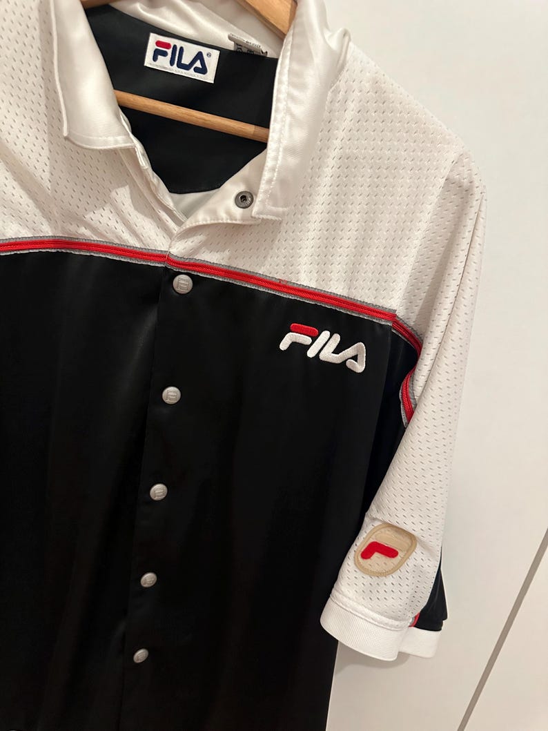 Peut inclure: Chemise boutonn&eacute;e Fila noire et blanche avec garniture rouge. La chemise pr&eacute;sente le logo Fila sur la poitrine et un &eacute;cusson sur la manche. La chemise a un haut en mesh blanc et une partie inf&eacute;rieure noire.
