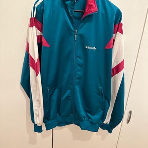 Könnte beinhalten: Türkisfarbene Trainingsjacke mit weißen und magentafarbenen Akzenten. Die Jacke hat einen durchgehenden Reißverschluss, einen magentafarbenen Kragen und das Adidas-Logo auf der Brust. Die Ärmel haben weiße Streifen mit magentafarbenen Details. Ein klassischer Sportstil.