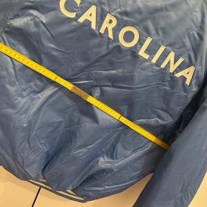 May include: A light blue jacket with the word "CAROLINA" in white block letters across the back. A yellow measuring tape is laid across the jacket, showing measurements in inches.