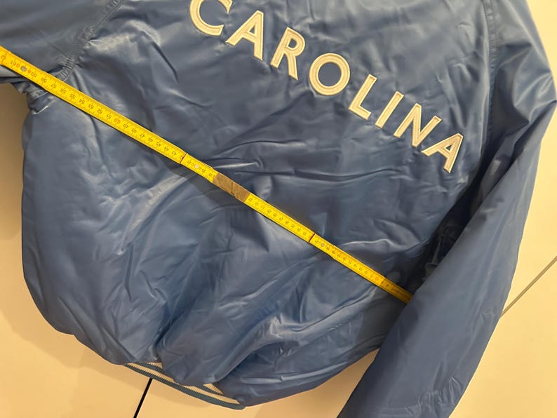 May include: A light blue jacket with the word "CAROLINA" in white block letters across the back. A yellow measuring tape is laid across the jacket, showing measurements in inches.