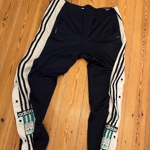 Op de afbeelding: Marineblauwe Adidas trainingsbroek met witte strepen langs de zijkanten. De broek heeft een drukknoopsluiting op de onderbenen en het Adidas-logo aan de zijkant. De broek is gemaakt van een glad, sportief materiaal.