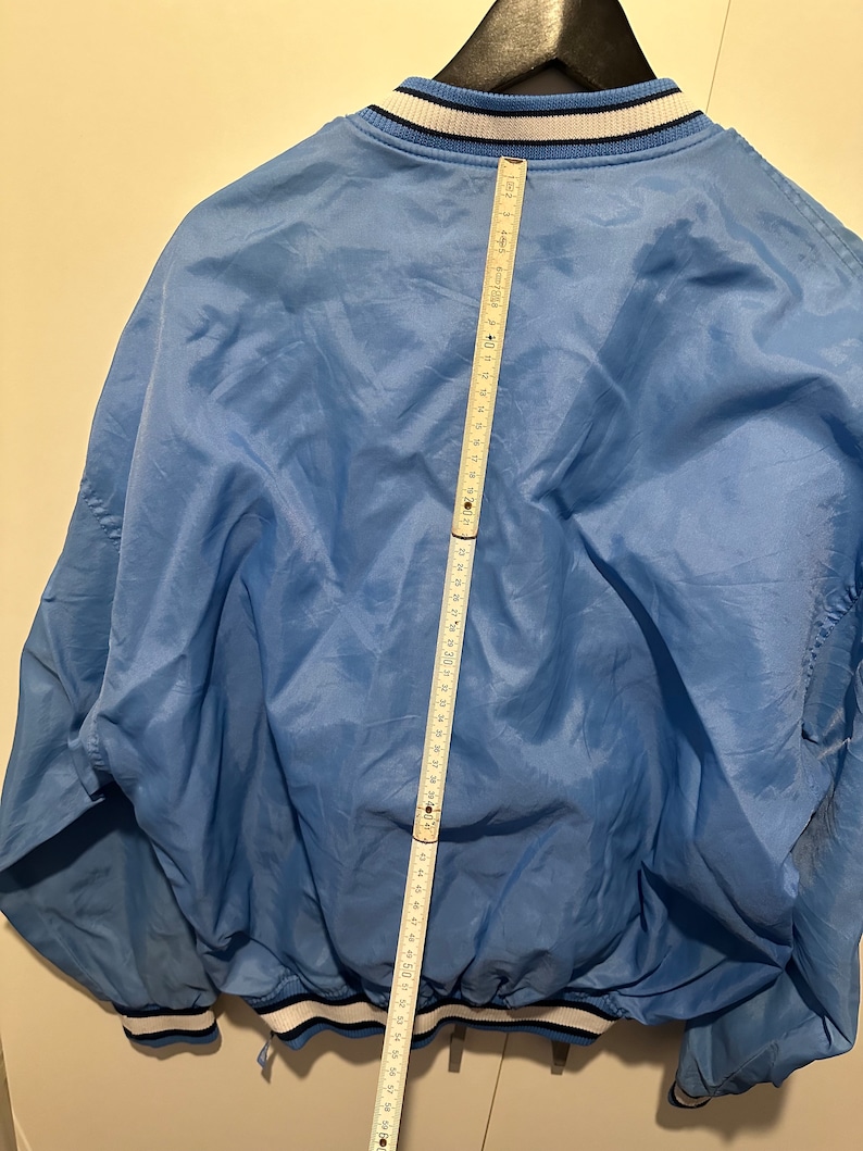 K&ouml;nnte beinhalten: Eine hellblaue Jacke mit wei&szlig;en und schwarzen Einfassungen an Kragen, &Auml;rmelb&uuml;ndchen und Saum. Die Jacke hat einen Rei&szlig;verschluss und einen Kordelzug in der Taille. Die Jacke wird mit einem Ma&szlig;band gemessen und zeigt eine L&auml;nge von 147 cm.