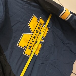 May include: Navy blue and black jacket with the word "MICHIGAN" in yellow block letters. The jacket has a yellow and white striped cuff. A yellow measuring tape is laid across the jacket.
