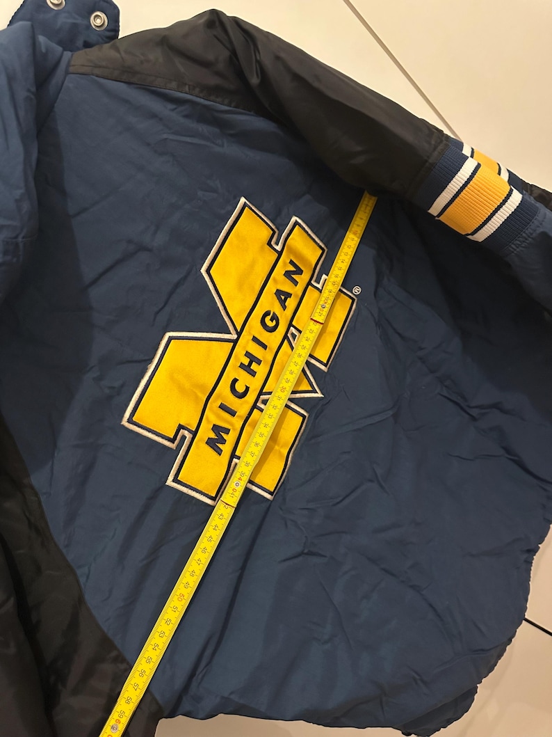 May include: Navy blue and black jacket with the word "MICHIGAN" in yellow block letters. The jacket has a yellow and white striped cuff. A yellow measuring tape is laid across the jacket.